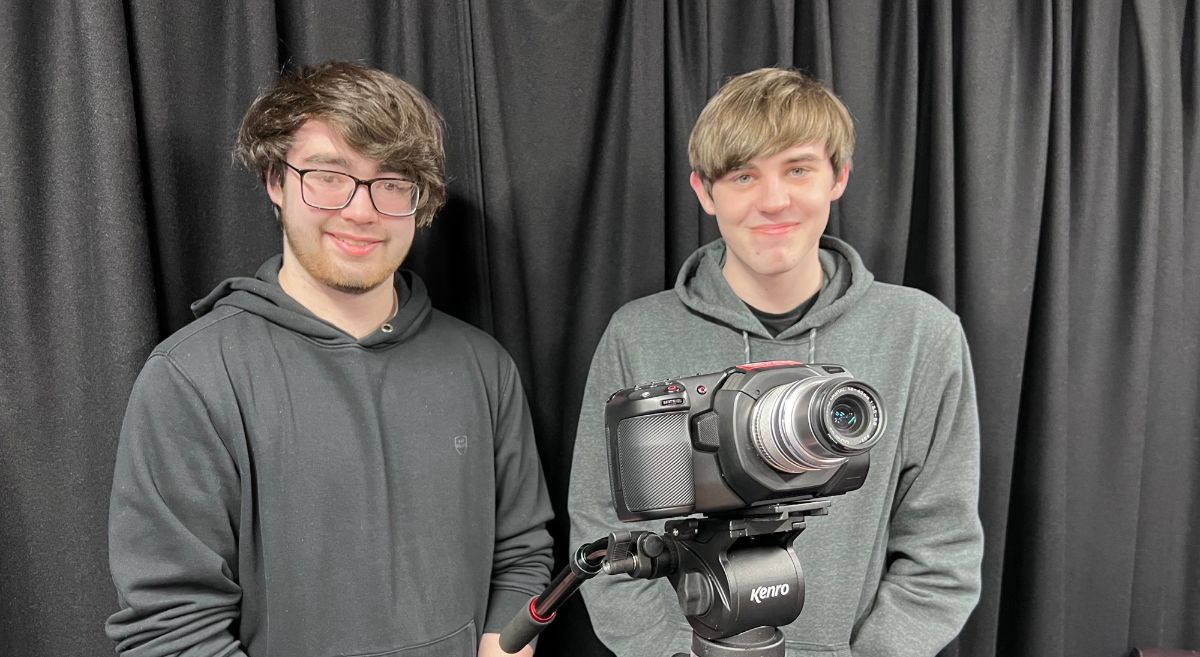 Two male students dress in black hoodies standing behind a camera on a tripod with black stage curtain in background